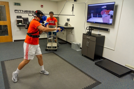 college student wearing a virtual reality headset and throwing a punch. A T V screen shows a simulation of a boxing ring.