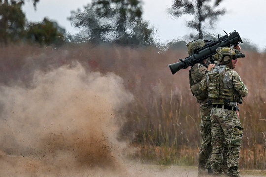 two American soldiers standing next to each other, one with a rocket launcher on his shoulder. There is debris and heat waves in the air behind them from a recent blast.