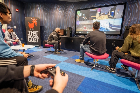 five people sitting on couches and chairs in a room playing a video game on a large screen.