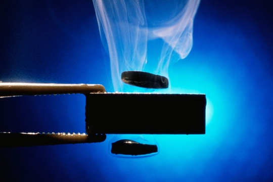 a closeup of tweezers holding a black disc with two smaller black discs hovering above and below it, with smoke rising into the air.