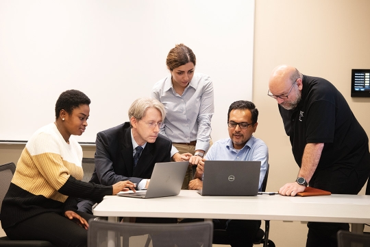 five students and faculty crowded around a table looking at two laptop computers.