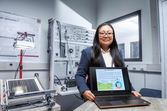 Bing Yan is shown working in a lab holding a laptop that is facing the camera showing a slide from a presentation deck