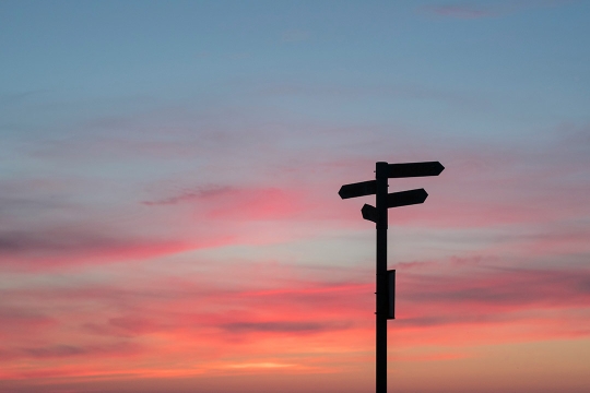 a wayfinding post pointing in different directions appears in front of a multicolor sunset in the sky