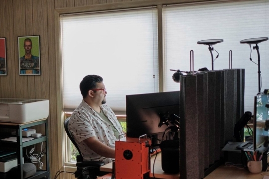 Juan Carlos Noguera appears sitting at a desk in his home with a window behind him