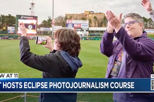 a child and an adult are seen at a ballfield photgraphing the sky during the eclipse.
