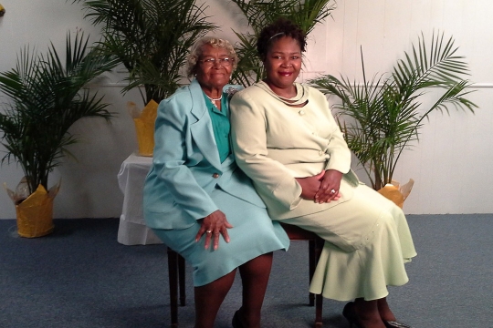 a black woman and her adult daughter sit on a bench with various plants behind them.