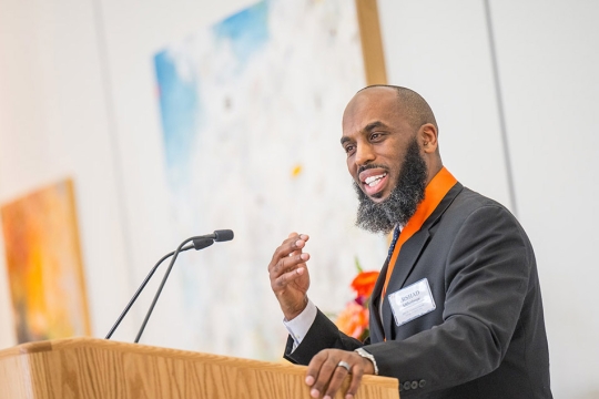 Irshad Altheimer appears at a podium in a black suit speaking to a crowd of people.