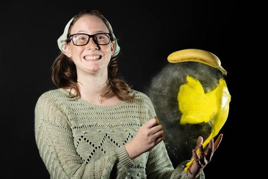 A person wearing glasses and a knit sweater smiles while holding a yellow balloon that has burst into a splash of powder. A banana floats above the ruptured balloon, all against a black background.