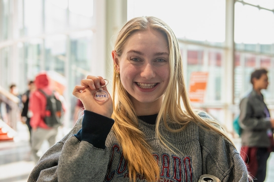 a young blonde student wearing a gray sweatshirt smiles and holds up a round clear keychain that says VOTE.
