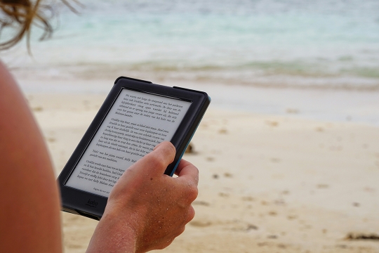 hands hold an ereader while relaxing on a beach.
