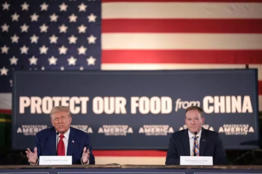 Donald Trump and another adult man sit at a table with an american flag and a poster saying Protect our Food from China displayed behind them.