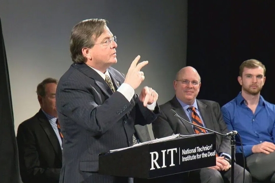 Gerry Buckley appears on a stage, signing to the audience.