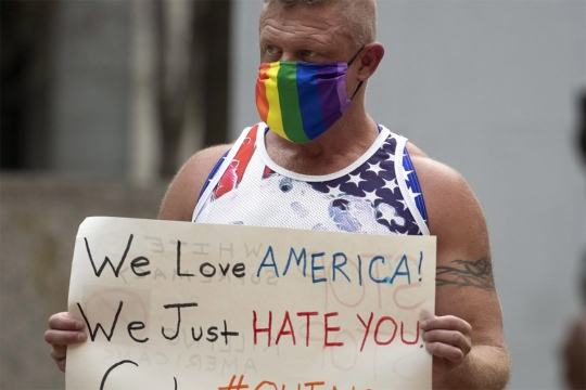 an older man in a rainbow face mask holds a sign  saying we love america we just hate you.