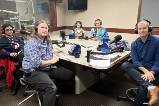 a group of people sit around a table wearing headsets ready for a radio show.
