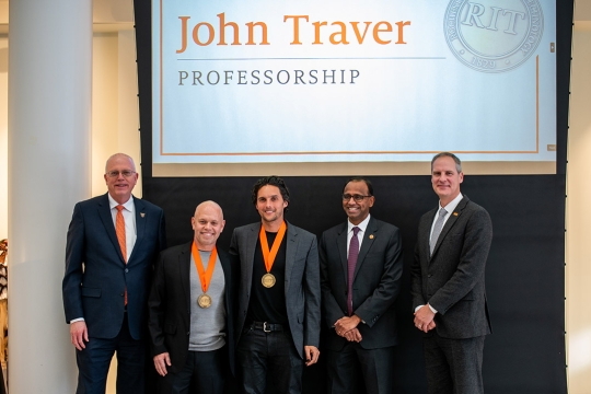 Five professionally dressed men stand together at an event, two wearing gold medals on orange ribbons. A large screen behind them displays 'John Traver Professorship' along with the RIT logo.