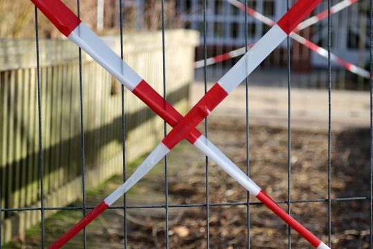 Red and white caution tape in an X shape on a metal fence, blocking access to a closed-off outdoor area.