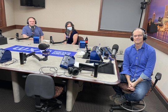 Three people sit at a table in a WXXI recording studio. The table has build in microphones in front of each chair and a WXXI logo in the center of the table. Three people sit around the table. Evan Dawson sits on the far right with two radio show guests sitting on the far left of the table. Thomas Warfield is not pictured.