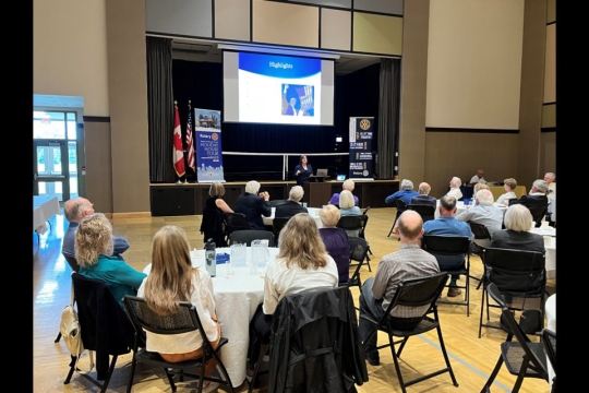 A room full of people seated at round tables, most of them seem to be older in age with white and gray hair. At the front of the room is a small stage and projector screen. In front of the stage, Sarah Burns stands in front of a podium with her laptop during her presentation.
