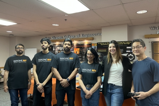 Six people pose for a photo in front of an academic poster. People are wearing shirts that read Imagine RIT.