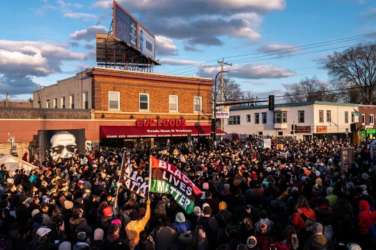 A photo by Joshua Rashaad McFadden showing a city street intersection filled with protestors. The street is no longer visible due to the large crowds. In the lower left corner of the image, two protestors wave flags that read Black Lives Matter. A mural of George Floyd painted on a local business is visible on the left side of the image.