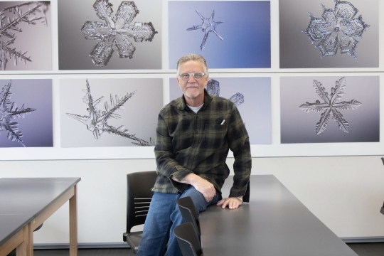 Man in green plaid flannel shirt leans on desk with eight up close photos of snowflakes behind him.