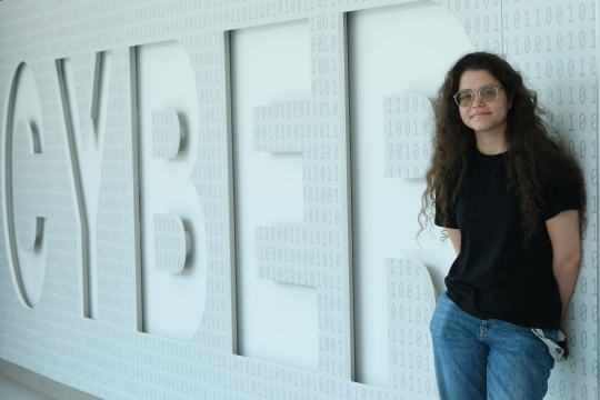 a young woman in a black shirt stands in front of a gray wall that reads CYBER in large letters.