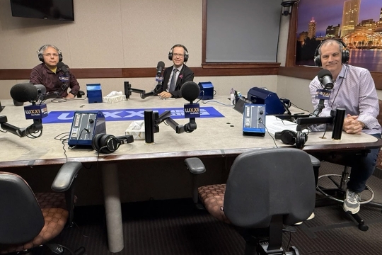 three men in headphones sit at a table with WXXI branded microphones.