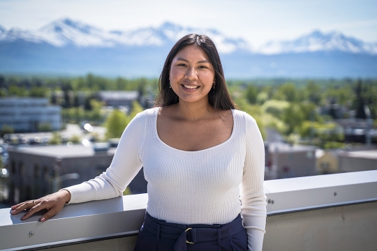 a college age woman in a shite shirt stands on a balcony with a town and mountains in the distance.