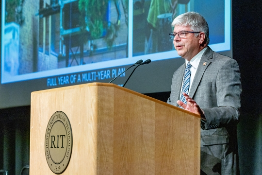 Bill Sanders stands at an R I T podium speaking to a crowd.