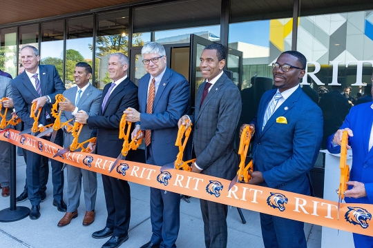Seven older men cut a large R I T printed ribbon in front of an art gallery