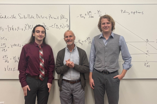 three men stand in front of a whiteboard in a classroom smiling at the camera.