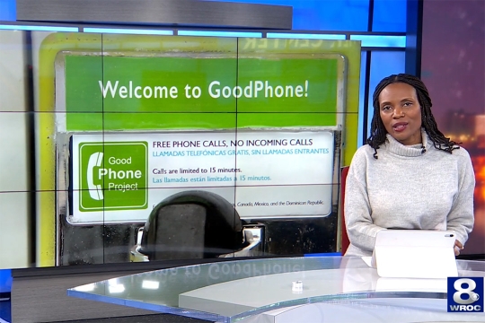 A news anchor sits at the newsdesk. To her left on a screen behind here is a close up image of one of the GoodPhone installations. The photo features a sign above the phone receiver that reads Welcome to GoodPhone and offers brief instructions in English and Spanish.