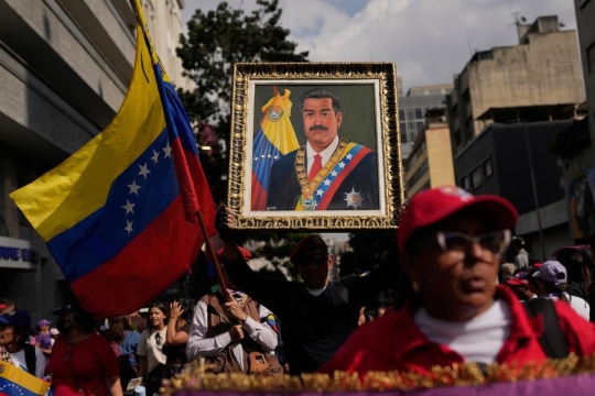 A group of people in Venezuela marching in the streets. The focus of the image is a large painted portrait of President Nicolas Maduro. To the left of the portrait someone waves a Venezuelan flag. Most of the people in the image are out of focus, with the portrait and flag being the focus of the image.