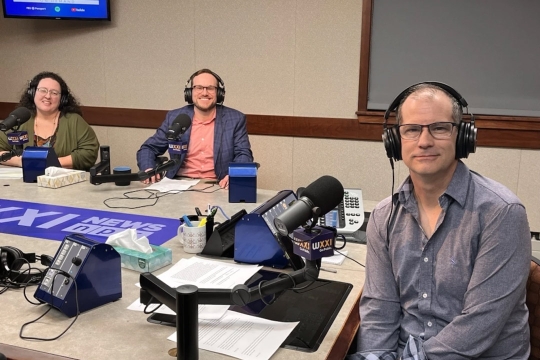 three people sit at a table wearing headphones and speaking in to microphones.