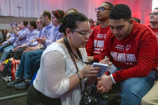 a woman speaks to a man while writing in a notebook.