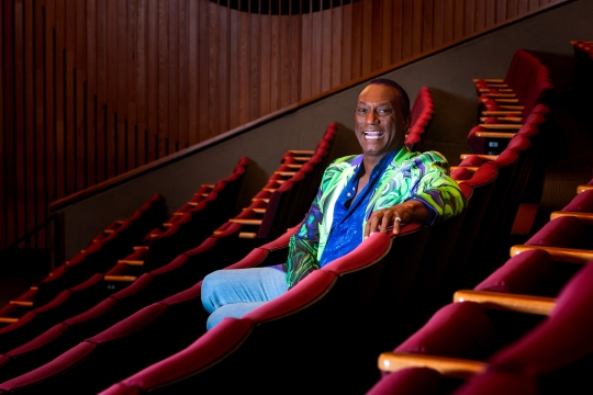 Thomas Warfield smiling sitting in an red chair in a theater wearing a colorful jacket and blue shirt and pants. 