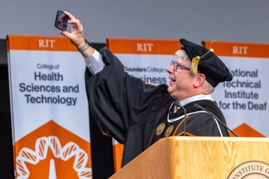 a man with white hair stands behind a podium in graduation regalia taking a selfie with a crowd.