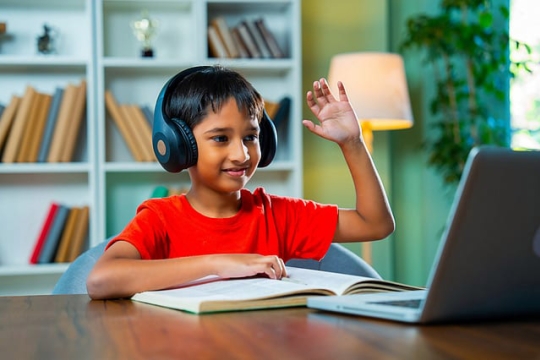 a child in a red tshirt holds his hand up while looking at a laptops screen. An open book is on a desk in front of him and he wears headphones.