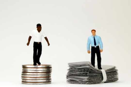 on the left, a plastic figuring of a black man stands on a pile of coins. On the right, a plastic figuring of a white man stands on a stack of paper money.