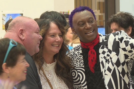 a man with close cut purple hair, a black and white blazer, and an orange scarf takes a selfie with a group of three people.