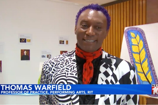 a man with close cut purple hair, a black and white blazer, and an orange scarf appear on screen behind a chyron with the name Thomas Warfield on it.