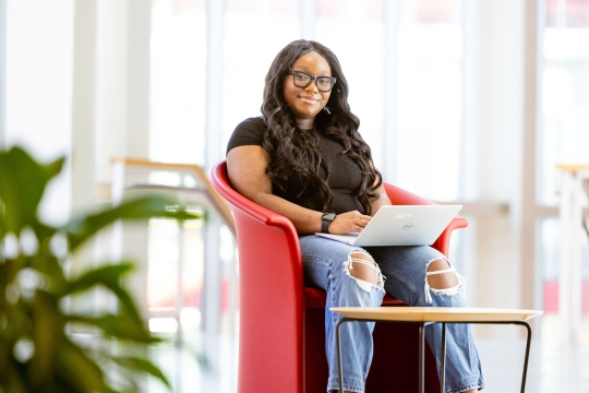 a college age woman with long brown hair sits in a brightly lit atrium in front of a laptop.