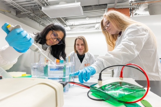three women in white lab coats use a large pipette and point at a liquid in a clear bowl
