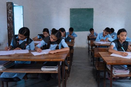 students sit working at desks in a rural classroom in india