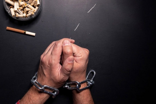 Hands bound by heavy chain sit on a black table with an ashtray of cigarette butts in the upper left corner.