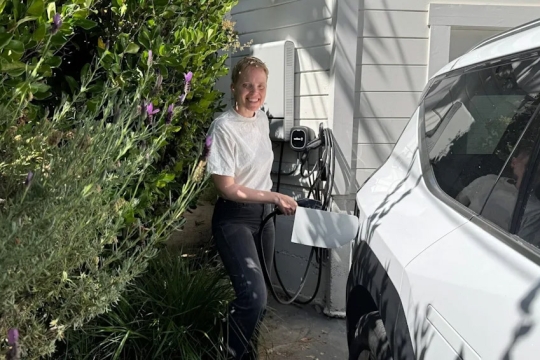 a woman in a white tshirt holds an E V charger next to a white car in a home driveway