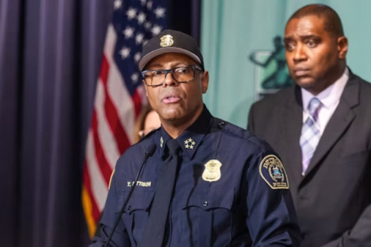 A Black man wearing a police uniform stands behind podium.