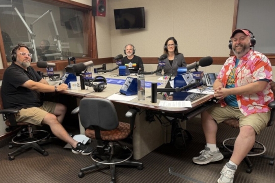 four people sit in a beige room in front of a table holding microphones.