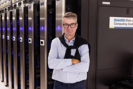 a man in a blue shirt with a navy blue sweater over his shoulders stands in front of a server farm