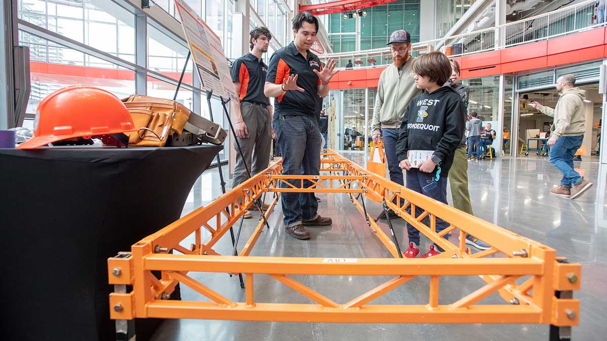 A student in a black polo shirt explains a structural engineering project to a young visitor at an exhibition event with a large orange bridge model.